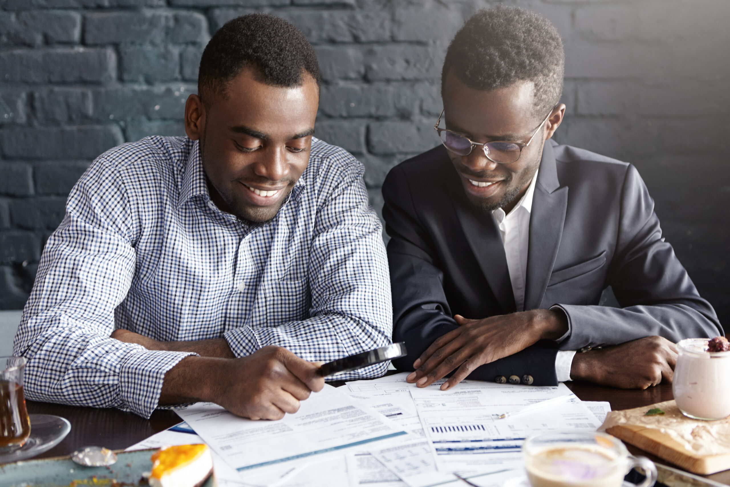 happy african office workers dressed in formal clothing having cheerful looks, studying and analyzing legal documents on table using magnifying glass while getting papers ready for meeting